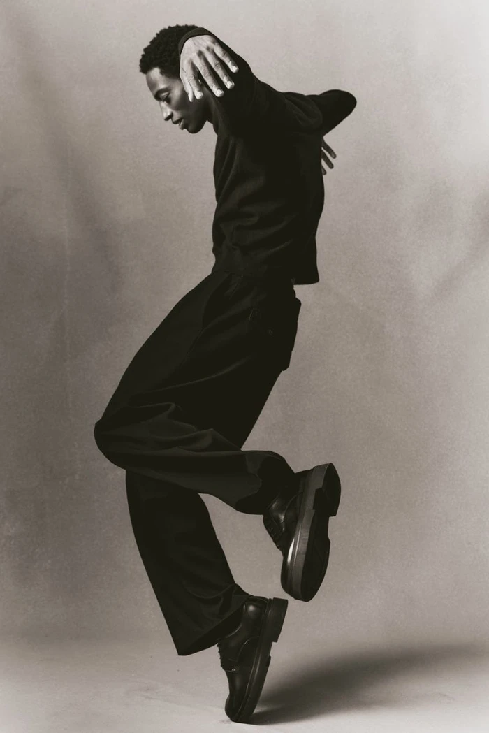 Black man wearing dark clothing and boots captured mid-jump against a neutral grey seamless studio background.