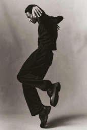 Black man wearing dark clothing and boots captured mid-jump against a neutral grey seamless studio background.