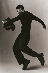 Monochrome fashion portrait of a black man wearing dark clothing and holding a dark handbag against a neutral background.