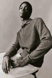 Man wearing a textured grey polo sweater and light trousers seated against a plain neutral backdrop.