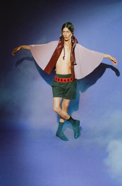 Adult male fashion model with braids poses in a modern runway look. Lavender cape and black shorts on a blue gradient backdrop.