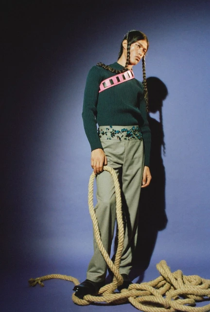 Male fashion model poses in a studio with a rope and dark green sweater, accessorized with a pink harness and floral belt, against a blue backdrop.