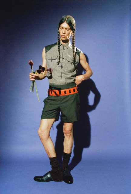 Full studio shot of a slender adult male with long braided hair, wearing a gray sleeveless shirt, black shorts, and holding flowers.