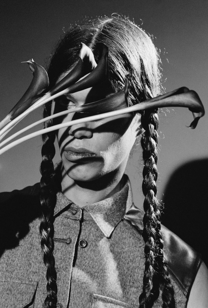 Monochrome portrait of woman with braided hair and a grey top. She is surrounded by calla lilies, casting shadows across her face.