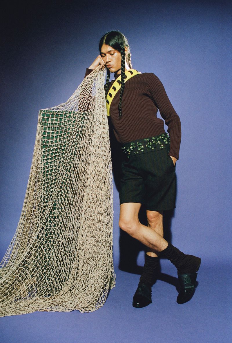 A person poses in front of a fishing net wearing a brown sweater, black shorts, socks, and loafers in a fashion editorial.