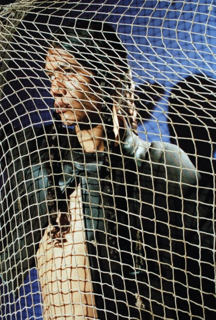 Adult female poses behind a tan and black net in a medium shot. The background is dark blue and out of focus, enhancing the model.
