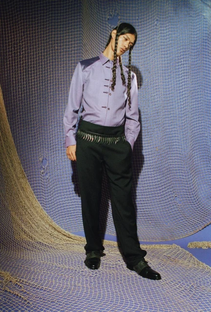 Full-length portrait of a person with braided hair wearing a lavender shirt and dark pants standing in front of a blue net background.