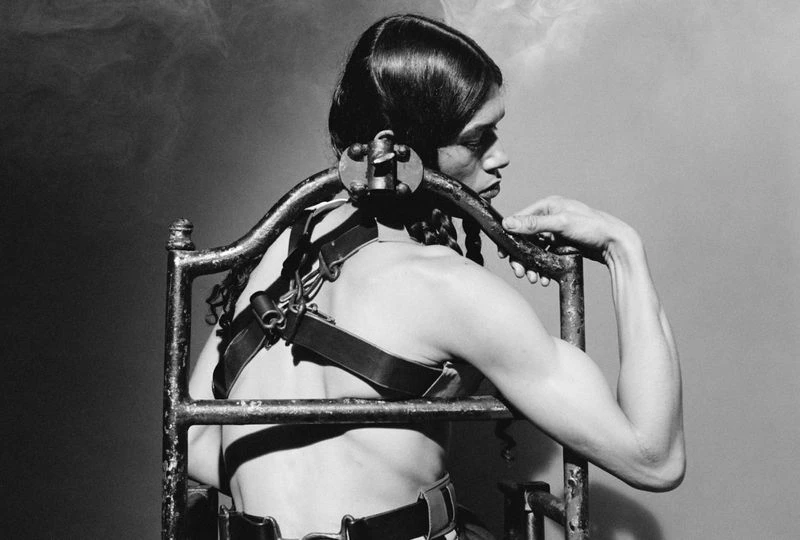 Back view of androgynous person with a shaved head and arm raised wearing a metal and leather restraint device in a smoky monochrome setting.