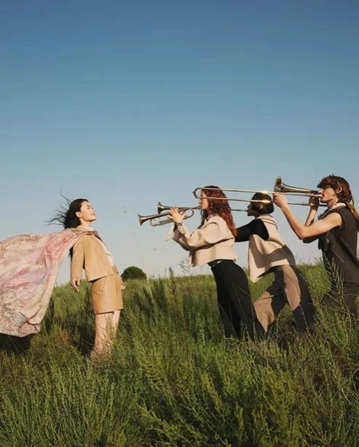 Group of musicians outdoors in a field, woman standing with patterned cape, other women playing trumpets, blue sky and tall grass.