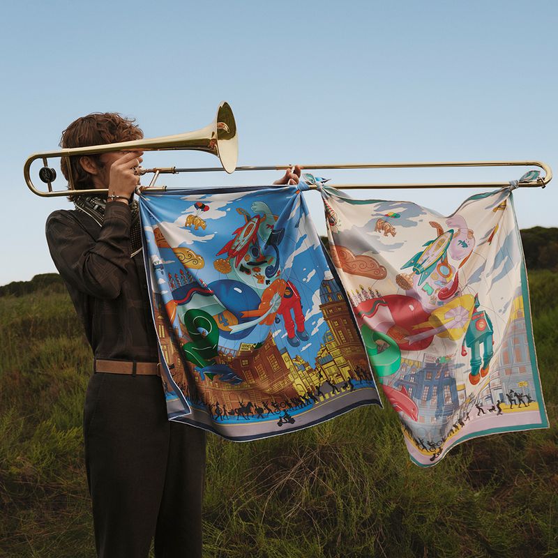 Young adult male plays a gold trombone, stands in a grassy field, and displays two illustrated textile art panels.