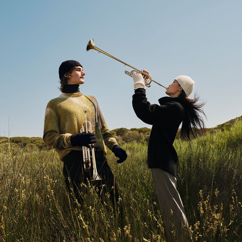 Two people, one male and one female, standing in a field playing trumpets under a clear sky. Wearing knit hats and stylish clothing.