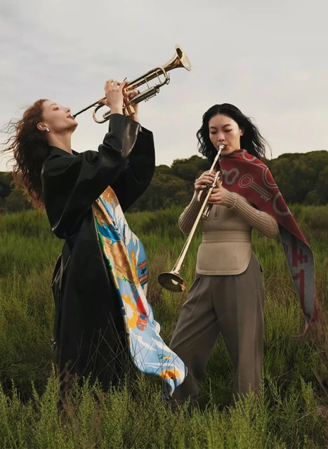 Two women in a grassy field play brass trumpets, one in a black coat with a colorful scarf, the other in neutral tones with a patterned shawl.