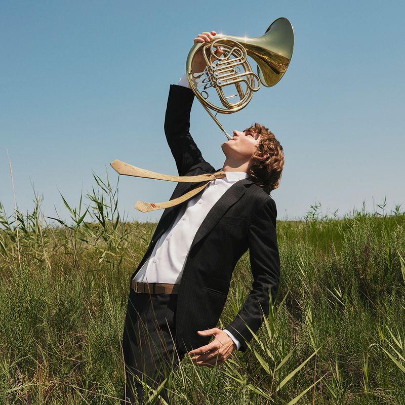 A man in a suit plays a French horn in a field of tall grass under a blue sky, creating a surreal, artistic image.