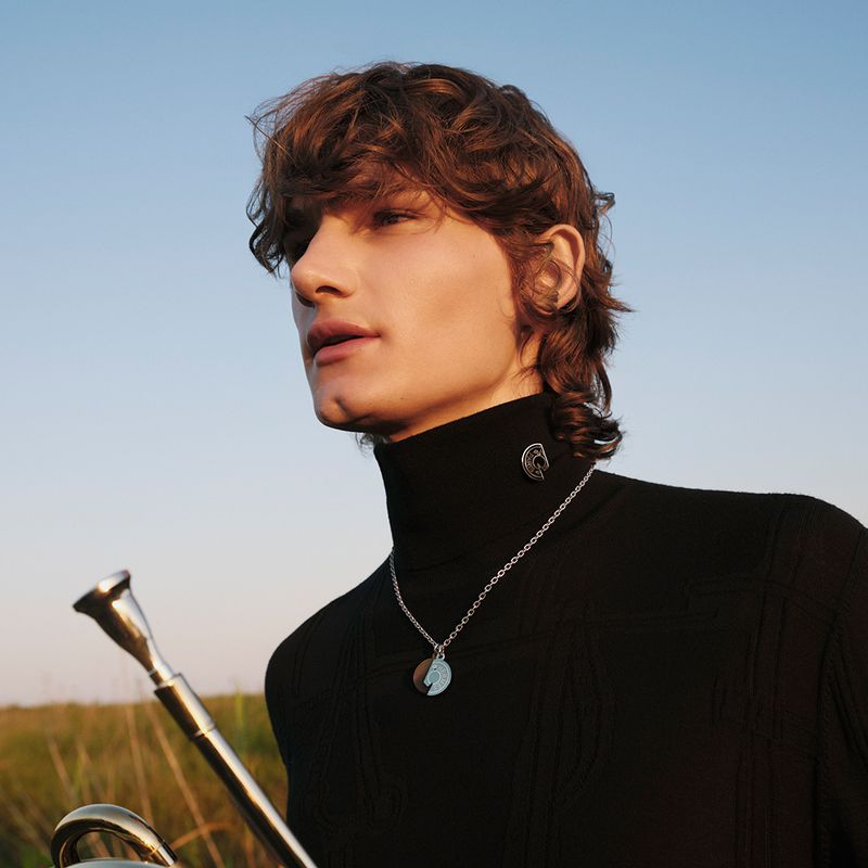 Portrait of a young man with brown hair wearing a black turtleneck and silver necklace, standing outside in a grassy field with a trumpet.