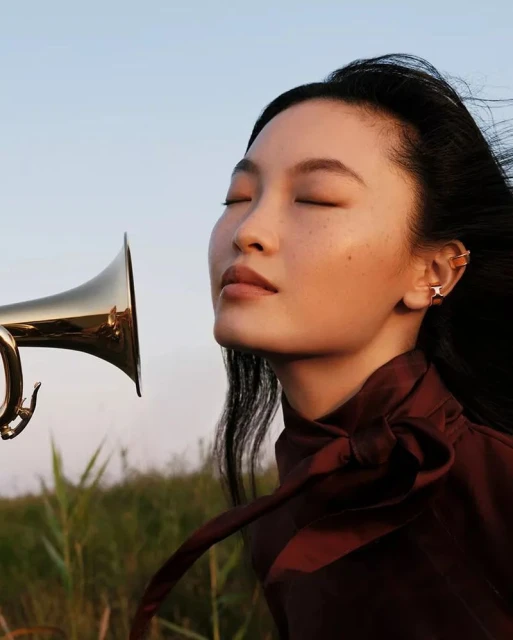 Portrait of an Asian female model with closed eyes next to a gold trumpet, outdoors in a field of tall grasses under a bright blue sky.