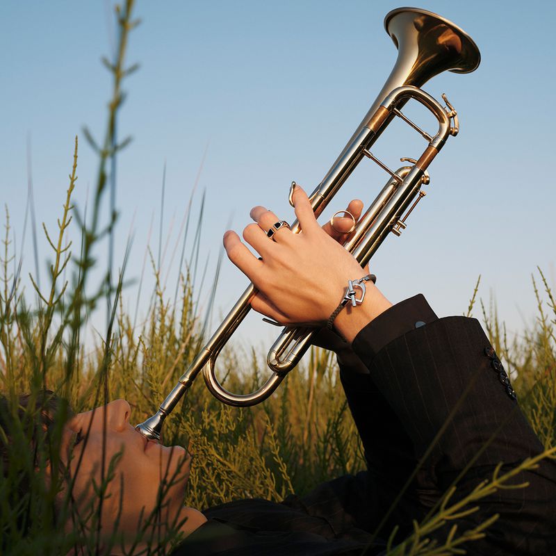 Person plays a trumpet outdoors, lying in tall grass against a clear sky, wearing a black suit jacket and wrist bracelet.