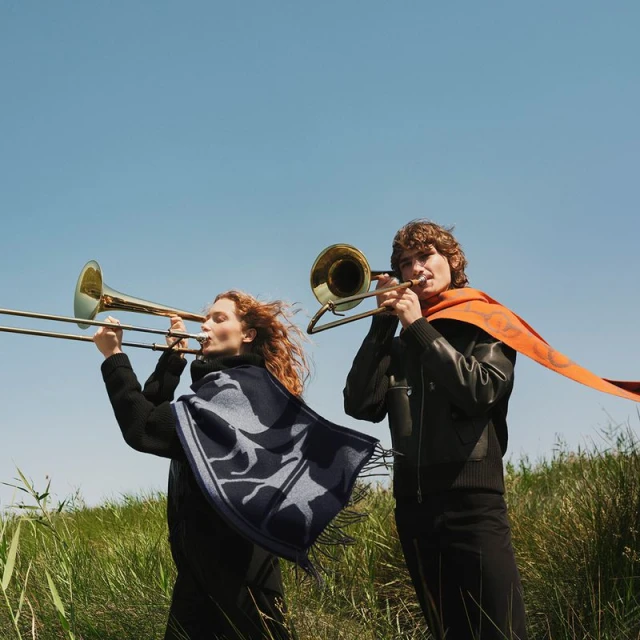 Two musicians play trombones outdoors in a field of tall grass under a clear blue sky, one wearing a patterned scarf and the other wearing a scarf.