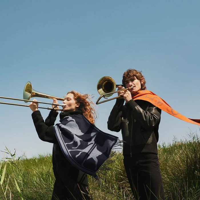 Two models playing trombones outdoors in a grassy field under a clear blue sky, wearing Hermes silk scarves.