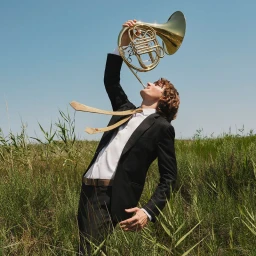Young man in a black suit holding up a golden French horn outdoors in tall green grass under a clear blue sky