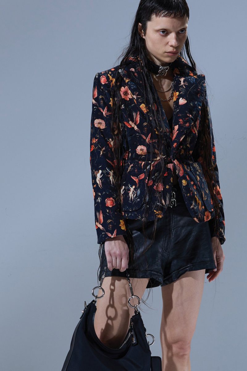 Young woman in a floral print blazer, leather shorts, and a black bag, posing indoors against a grey backdrop, showcasing fashion and style.