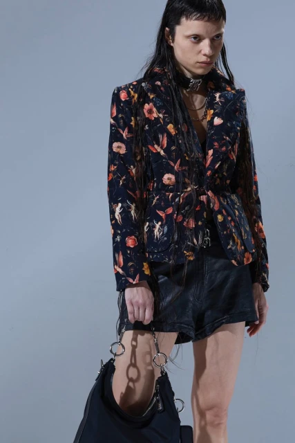 Young woman in a floral print blazer, leather shorts, and a black bag, posing indoors against a grey backdrop, showcasing fashion and style.