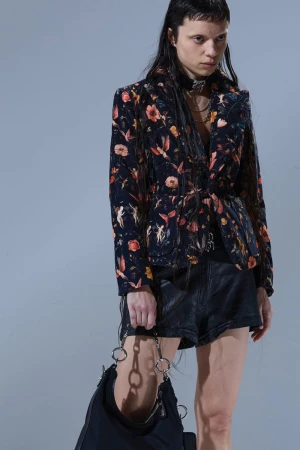 Young woman in a floral print blazer, leather shorts, and a black bag, posing indoors against a grey backdrop, showcasing fashion and style.
