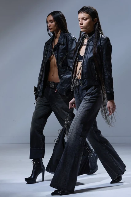 Two fashion models in black leather jackets and dark denim walk in a studio with neutral lighting. Fashion editorial, dark aesthetic.