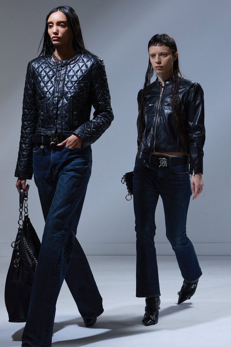 Two fashion models walk in black leather jackets and dark denim jeans. One carries a black leather chain bag in a studio setting.