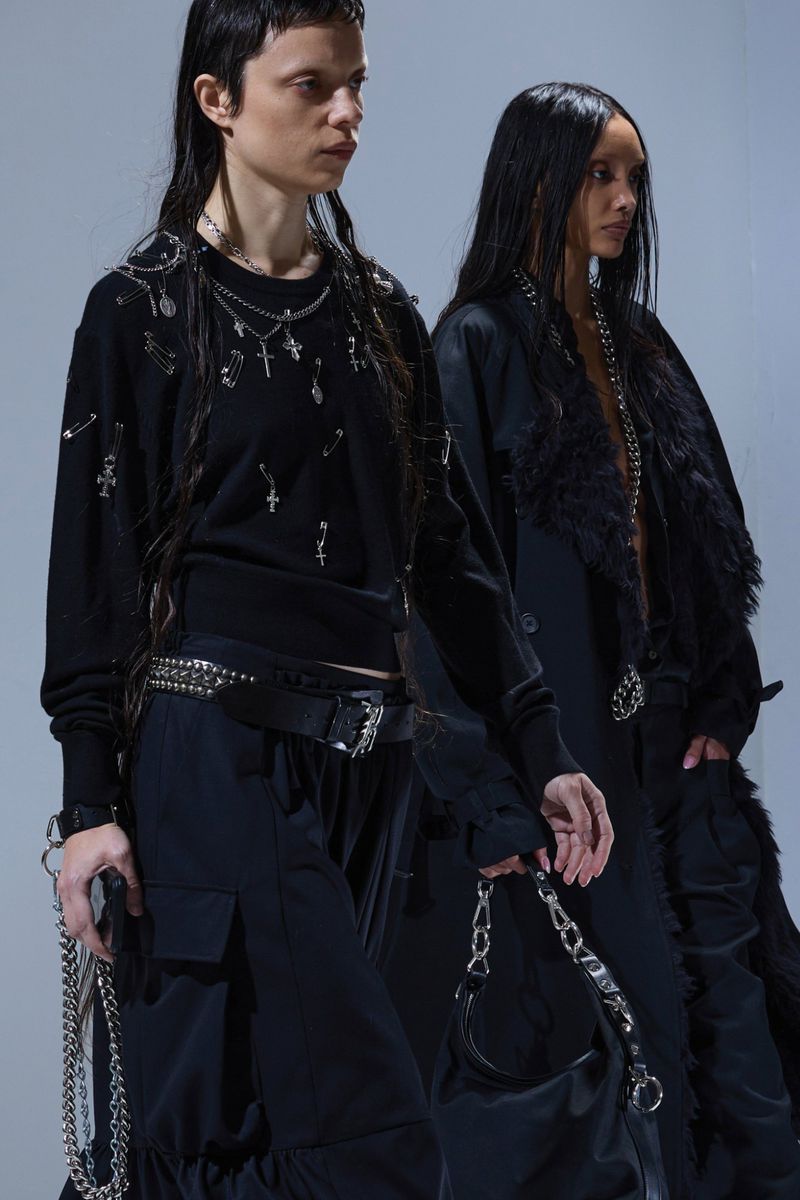 Two women in dark, edgy fashion stand side-by-side in a studio setting. Chains, crosses, and hardware add texture and detail to the scene.