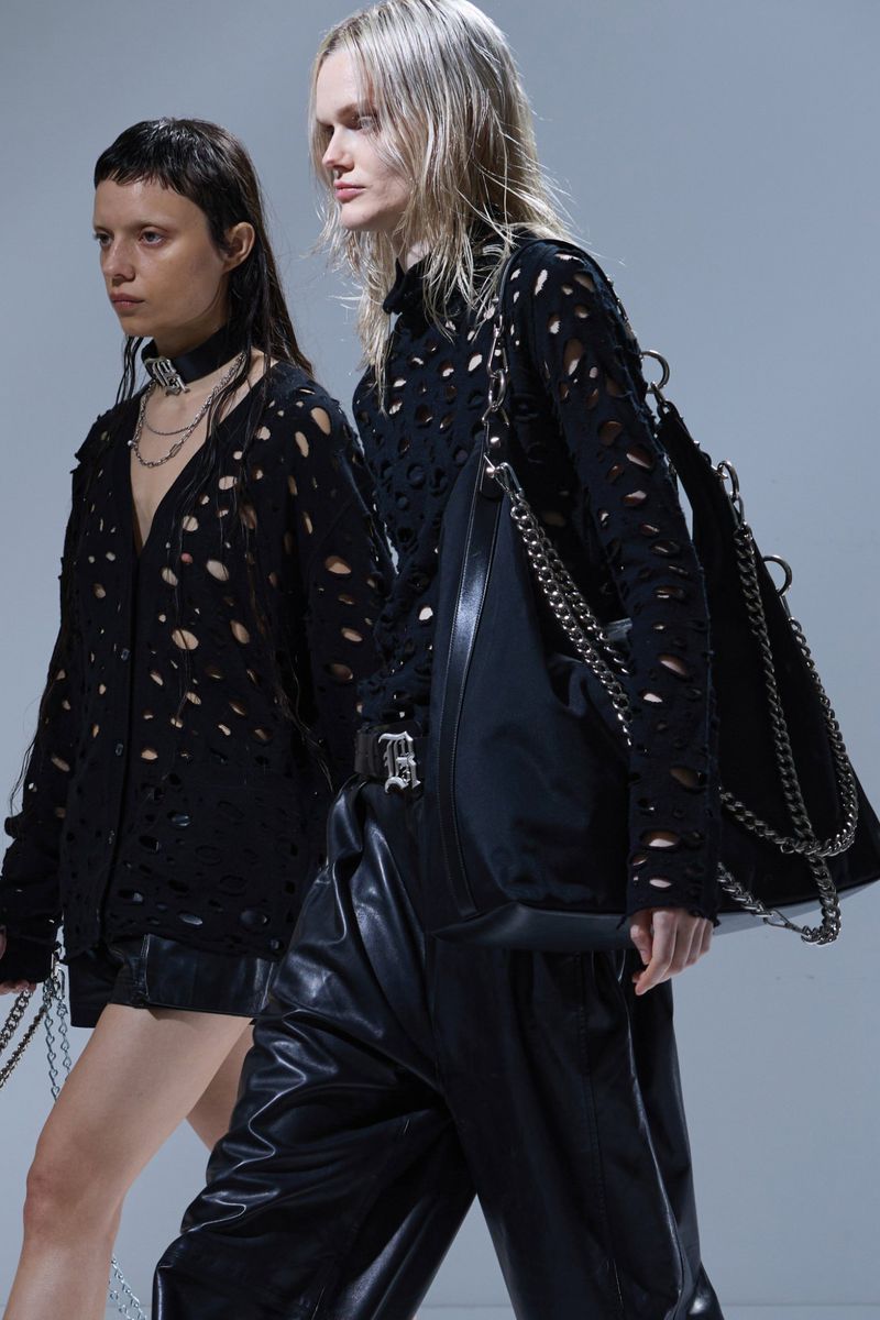 Two fashion models showcase black, distressed knitwear and leather pants against a light background. One carries a chain-strapped bag.