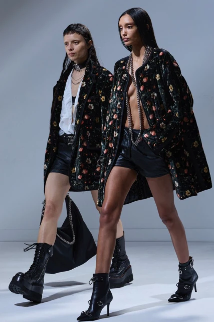 Two fashion models walk in a runway show wearing floral print jackets, black shorts, platform boots and carrying a handbag.