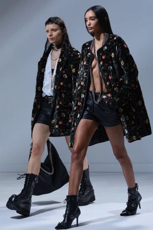 Two female models walking on a white studio backdrop wearing floral print jackets and black boots for R13 Fall 2025 Ready-To-Wear.