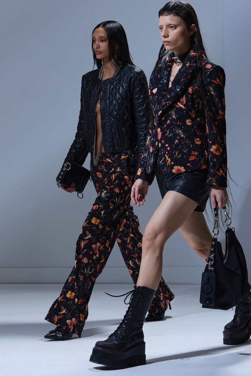 Two women walking in a fashion runway show. One is wearing a black jacket with floral pants and the other is in a floral jacket.