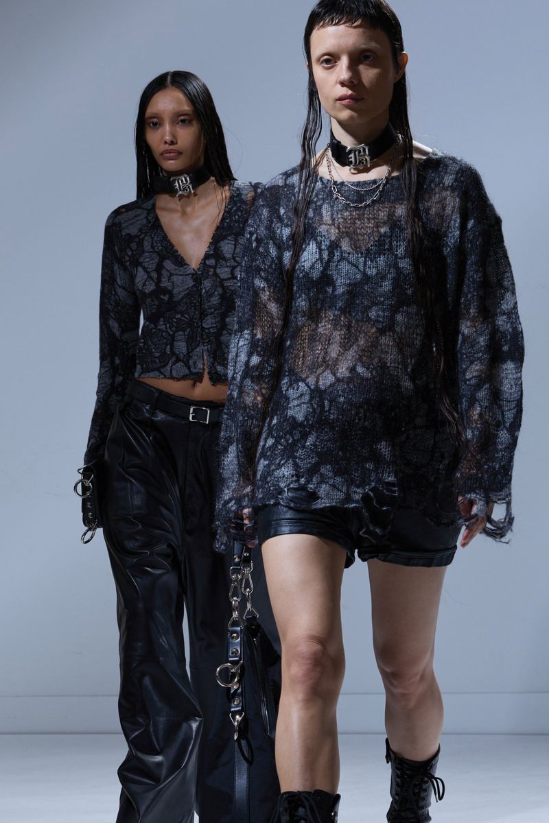 Two fashion models showcase dark apparel and accessories in a studio setting, featuring textured tops, leather pants, and edgy choker necklaces.