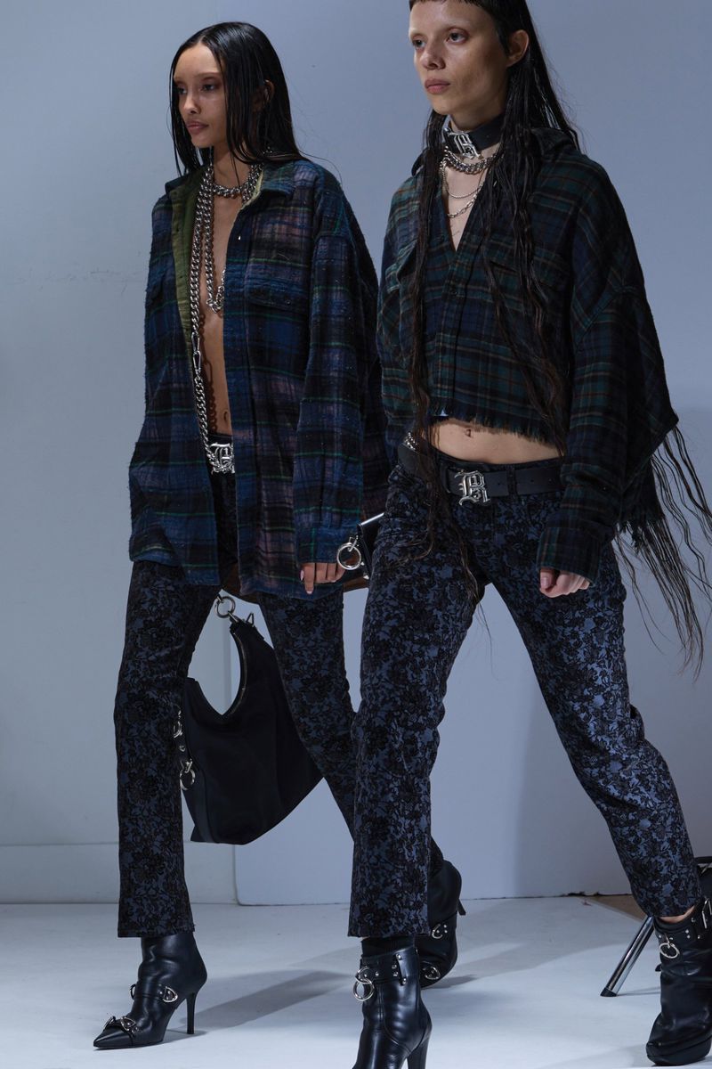 Two women wearing black, blue and green plaid shirts, patterned pants and black boots stand against a grey background in a studio shot.