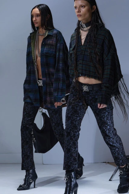 Two women wearing black, blue and green plaid shirts, patterned pants and black boots stand against a grey background in a studio shot.