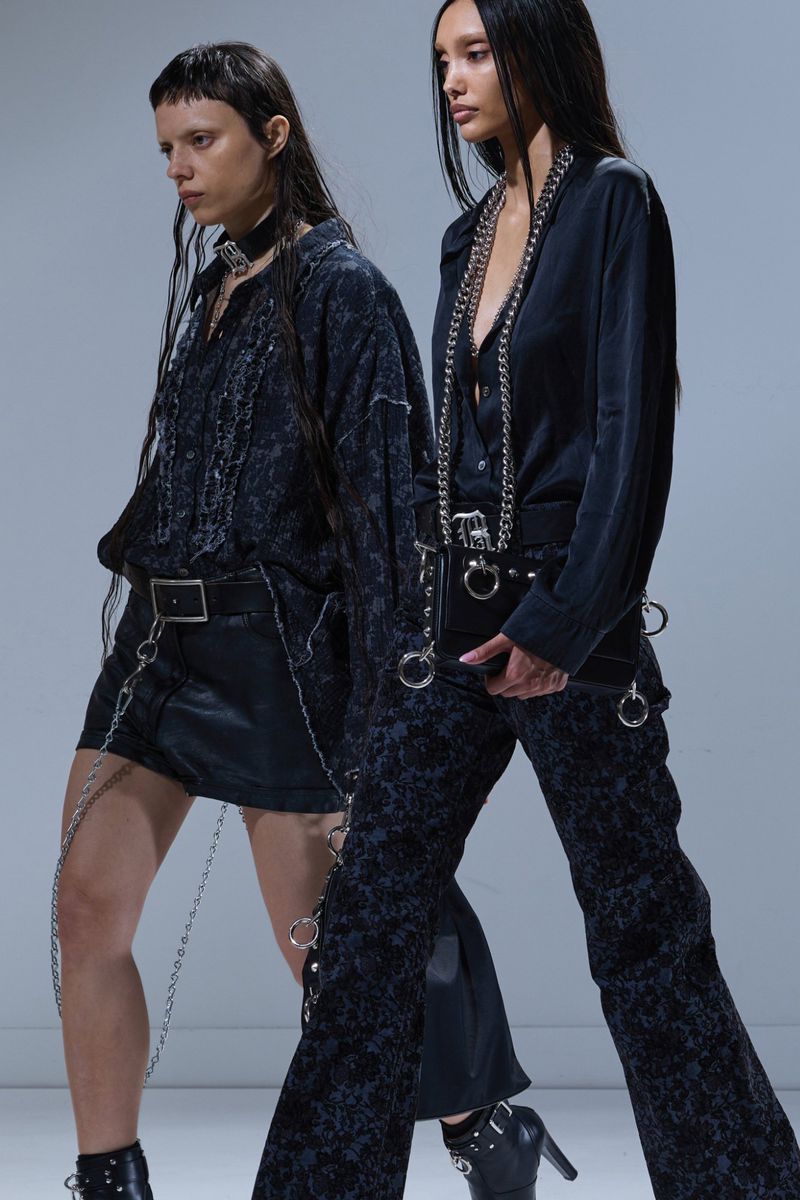 Two women in dark, textured clothing pose in a studio. One wears a mini-skirt and the other wears patterned pants with a matching jacket.