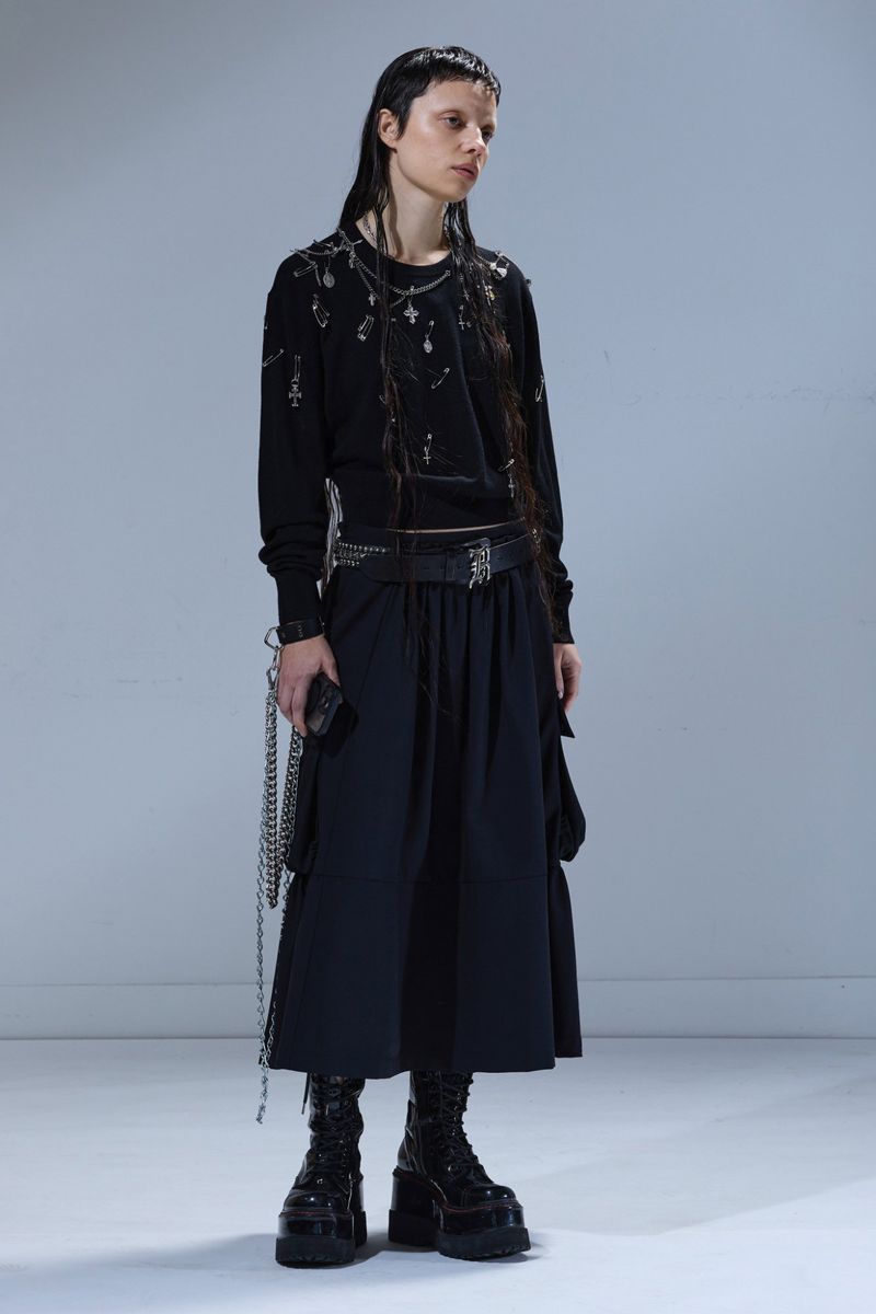 A slender young woman stands in a full studio shot, wearing a stylish black sweater, skirt, boots, and accessories in a neutral setting.