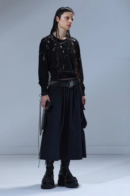 A slender young woman stands in a full studio shot, wearing a stylish black sweater, skirt, boots, and accessories in a neutral setting.