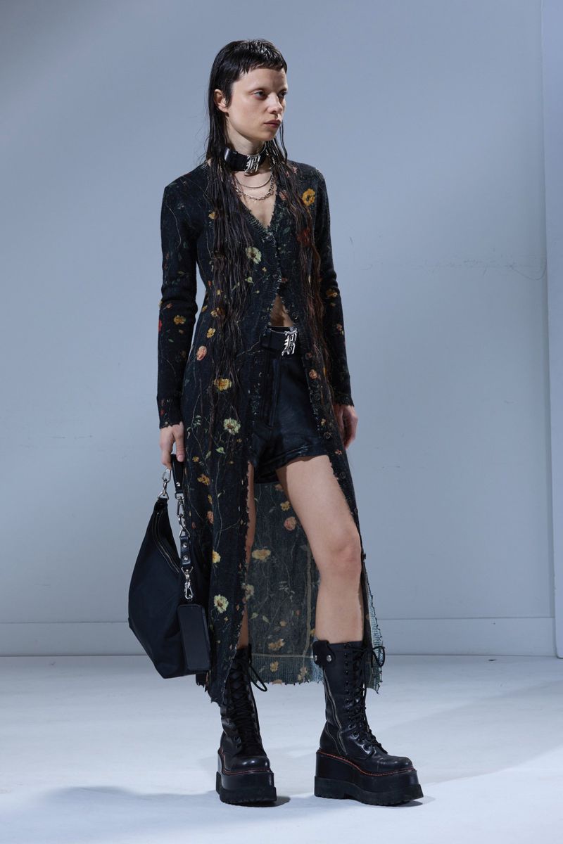 Model wearing a black floral cardigan and platform boots holding a black handbag against a white background