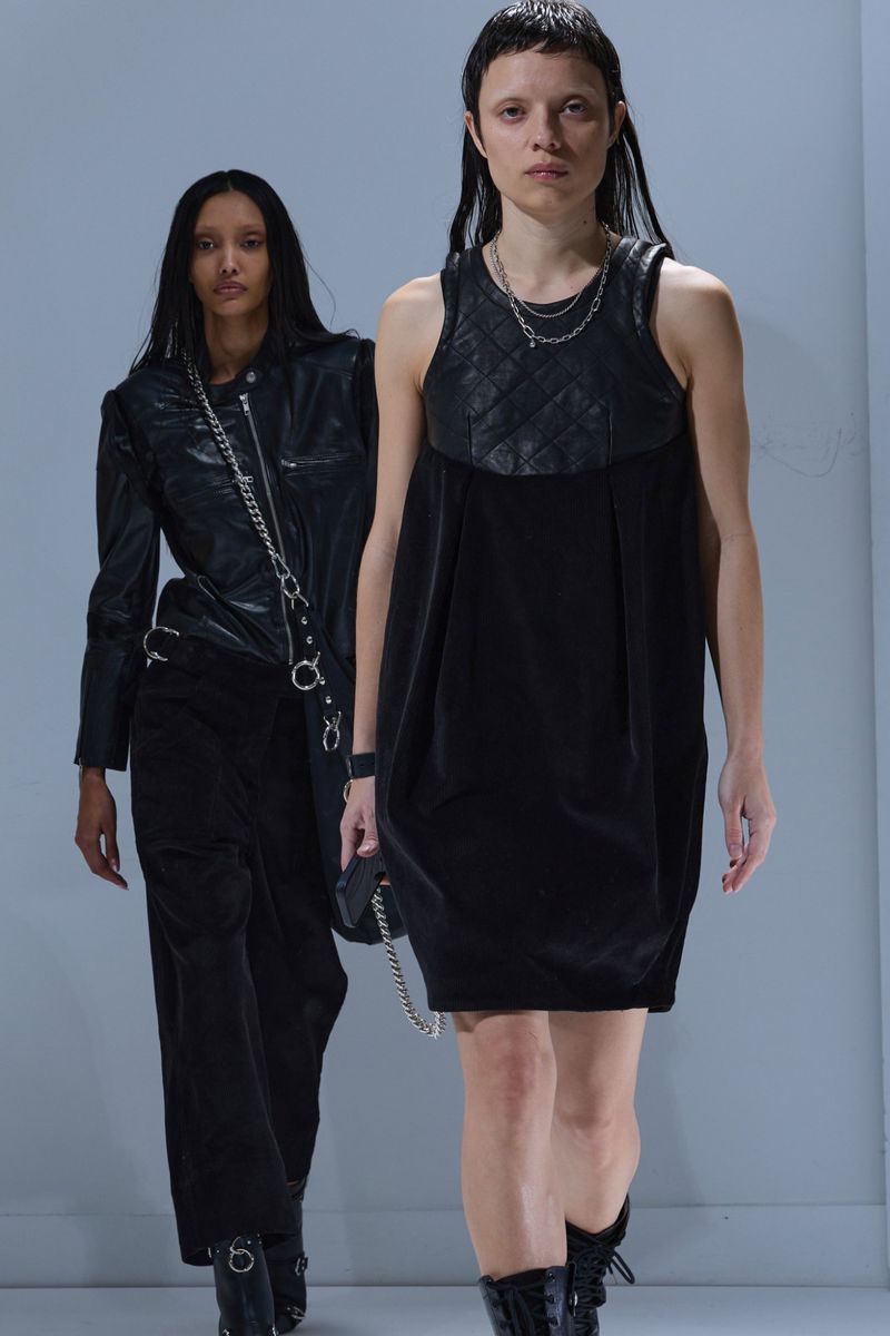 Two women wearing black clothing and boots on a runway. One woman wears a leather jacket, and the other wears a dress with a quilted top.