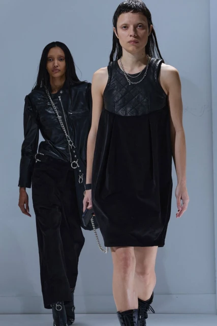 Two women wearing black clothing and boots on a runway. One woman wears a leather jacket, and the other wears a dress with a quilted top.