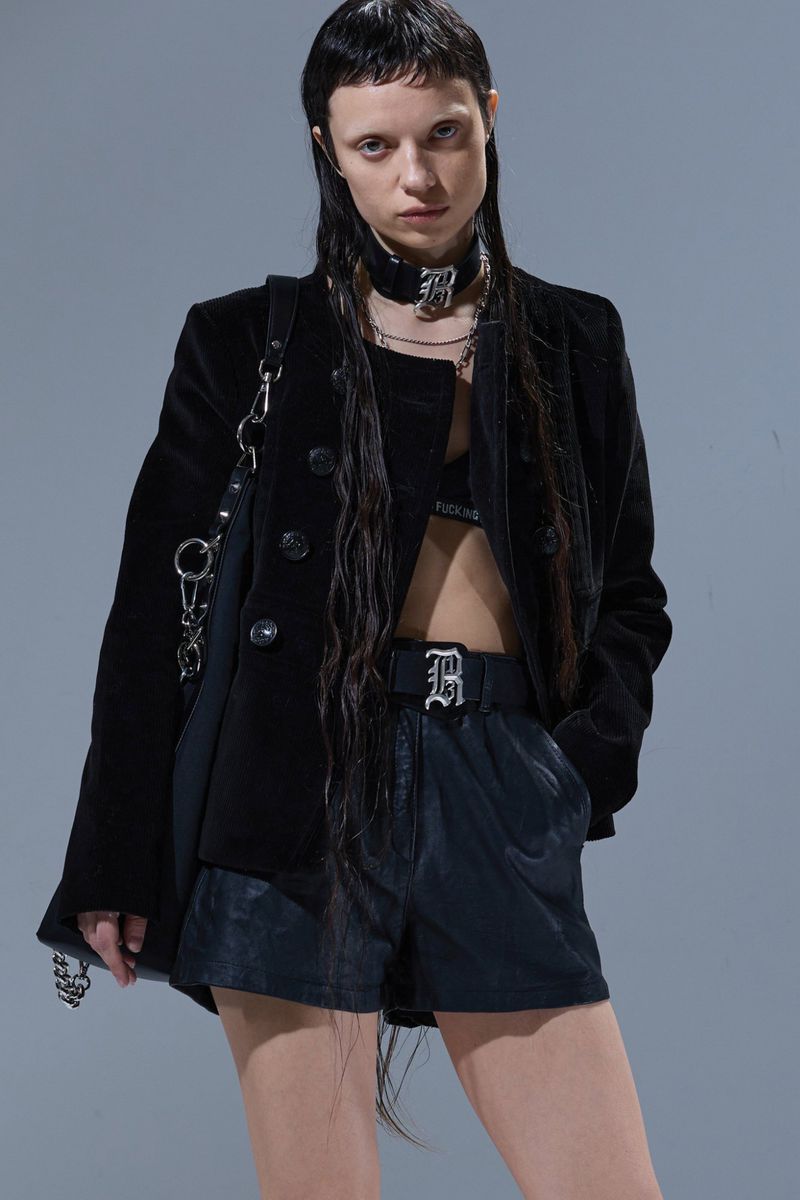 Woman in black blazer, bra top, shorts, and chain jewelry stands in a photo studio. Fashion shot with grey backdrop and soft lighting.