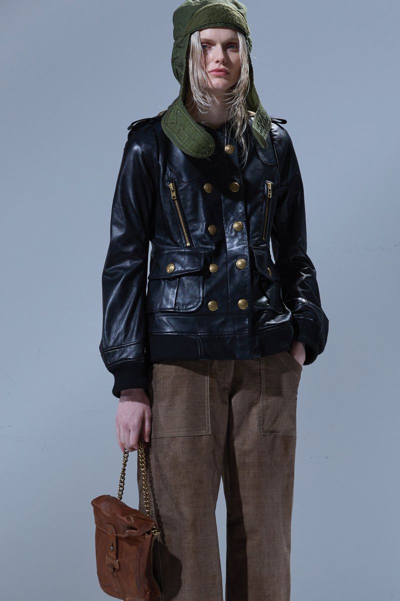 Fashionable young woman posing in a black leather jacket, brown pants, and green hat, holding a brown leather bag on a gray background.