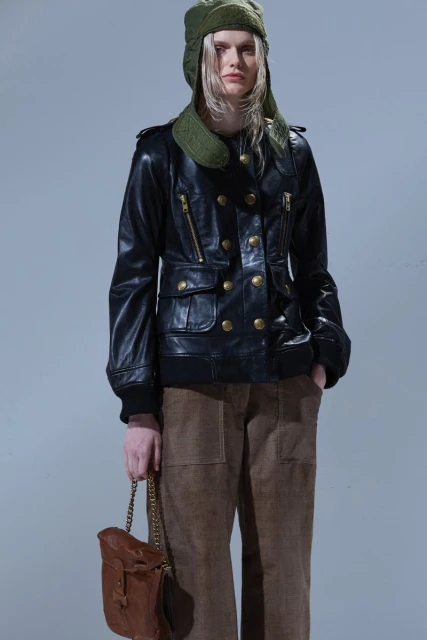 Fashionable young woman posing in a black leather jacket, brown pants, and green hat, holding a brown leather bag on a gray background.