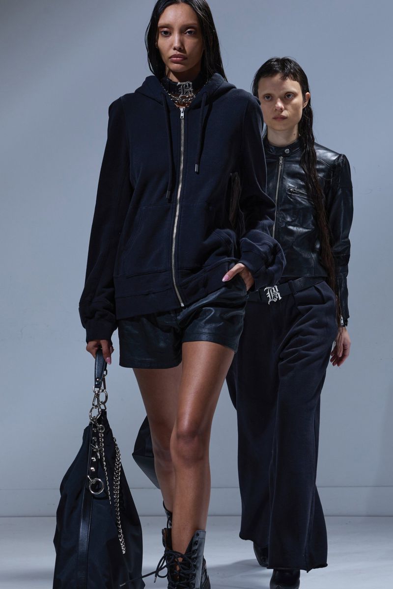 Two fashion models present dark apparel in a studio setting; one wears a zip hoodie, black shorts, and boots, while the other wears a leather jacket and pants.