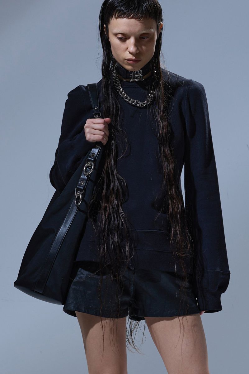 A woman in a black outfit poses with a black handbag in a studio. She is wearing a chain choker and has long, wet hair.