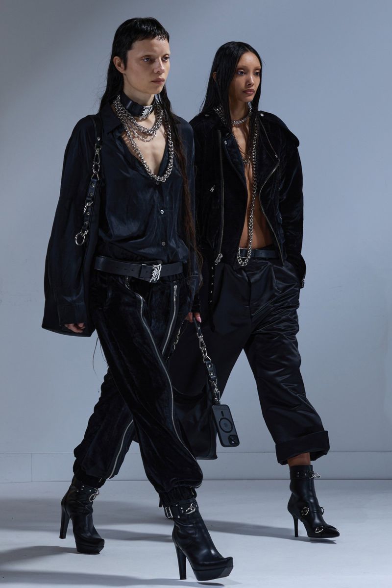 Two women in matching dark velvet outfits with edgy silver chain accessories are walking in a light grey studio setting.