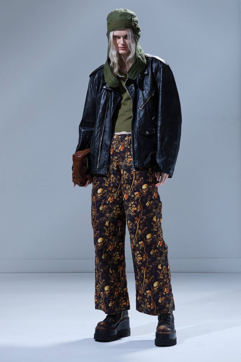 A full-length shot of a fair-skinned person with long blonde hair wearing a black jacket, floral pants, hat, and platform boots.
