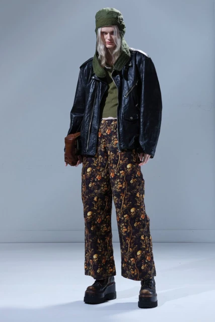 A full-length shot of a fair-skinned person with long blonde hair wearing a black jacket, floral pants, hat, and platform boots.